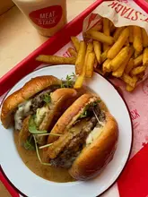 A top-down, high-angle shot of a messy, saucy burger meal from Meat:Stack, featuring a dry-aged steak burger with various toppings, served with fries and a shake on a branded red tray.
