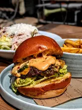 A high-quality, close-up food photography shot of a double smash burger with melted cheese and sauce, served with fries and a salad in the background. The image captures a casual, appetizing lunch setting, likely outdoors.