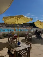 A bright, sunny, lifestyle-focused shot of an outdoor seating area at a lido, featuring yellow umbrellas and tables overlooking the pool.