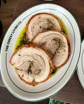 A top-down, overhead shot of three slices of porchetta served on a branded plate with a green herb oil, likely part of a roast dinner offering.
