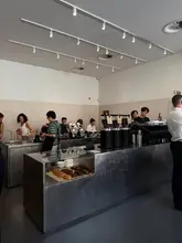 A bright, industrial-style coffee shop interior featuring a sleek metal counter and a display of pastries, capturing a candid moment of customers and staff.
