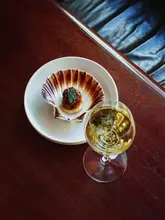 A high-angle, close-up shot of a single Isle of Mull scallop served in its shell with XO sauce and a shiso leaf, accompanied by a glass of white wine on a dark wooden table.