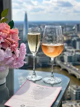A high-quality, bright shot of two glasses of wine on a glass table, set against a stunning panoramic view of the London skyline, including The Shard. The scene is styled with a bouquet of pink flowers and a menu, creating an inviting, upscale atmosphere.