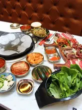 A high-angle, overhead shot of a Korean BBQ spread featuring various banchan, raw marinated meats, lettuce wraps, and a grill. The table is well-laid out, showcasing the variety of the meal.
