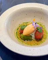 A high-quality, overhead shot of a beautifully plated dish from a tasting menu, featuring a vibrant orange sorbet, fresh tomatoes, and courgette, garnished with edible flowers. The presentation is elegant and artistic, typical of fine dining, set against a dark, minimalist background that allows the colours of the food to pop.