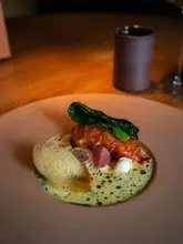 A high-quality, professional-grade food photograph showcasing a beautifully plated lobster dish at Ikoyi. The lighting is moody and focused, highlighting the textures of the lobster, the vibrant greens, and the glossy sauce. The composition is clean and deliberate, emphasizing the fine-dining nature of the venue.