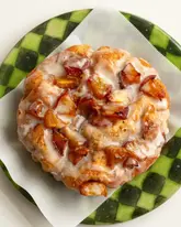 A high-quality, top-down food photography shot of a mini peach and cinnamon butter focaccia, glazed with vanilla icing, served on a green checkered plate.