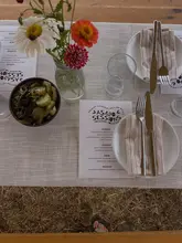 A top-down, aesthetically pleasing shot of a rustic, farm-to-table dining setup for an 'Asado Sessions' event. The image features a neatly laid table with a printed menu, fresh flowers, and a bowl of pickles, set against a backdrop of dry grass, emphasizing the outdoor, nature-focused setting.