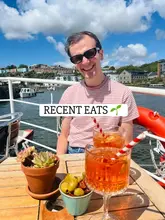 A casual, sun-drenched lifestyle shot featuring a person enjoying drinks and snacks at an outdoor, waterside venue. The image captures a relaxed, summery atmosphere, though the photography suffers from harsh lighting conditions.