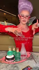 A promotional video still featuring a drag performer dressed as a fairy godmother, presenting themed green cocktails in a dark, atmospheric venue, likely for a Shrek-themed event at the Edinburgh Fringe.