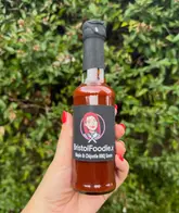 A clear, well-lit product shot of a 'BristolFoodie.x' branded Maple & Chipotle BBQ sauce bottle held in a hand against a soft-focus green background, promoting a collaboration with a local producer.
