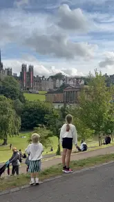 A candid, outdoor lifestyle shot taken in Edinburgh, likely Princes Street Gardens, capturing children looking out towards the city skyline during the Edinburgh Fringe festival. The image serves as a travel and family-oriented snapshot, documenting a day trip.