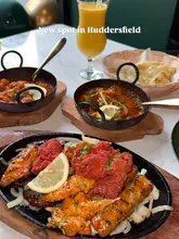 A table spread shot at an Indian restaurant in Huddersfield, featuring a sizzling platter in the foreground, two curry dishes, a mango lassi, and a basket of naan.
