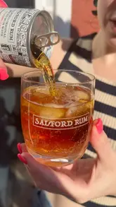 A bright, sunlit close-up shot capturing a canned rum and cola drink being poured into a glass filled with ice. The image focuses on the refreshing nature of the beverage, utilizing natural light to highlight the liquid and the condensation on the glass.