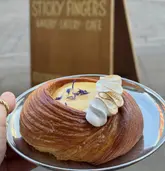 A close-up, eye-level shot of a lemon curd pastry held in a hand, featuring a swirl of meringue and edible flowers. The background shows a blurred wooden sign for the venue, providing clear branding context.