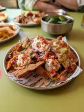 A top-down, overhead shot of a plate of 'naanchos' (naan nachos) served at a restaurant, with other dishes and a metal bowl visible in the background.