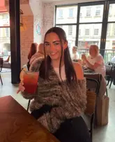 A portrait of a woman holding a cocktail in a casual bar or restaurant setting. The image captures a social moment, though the lighting is inconsistent with a distracting warm light spill on the subject's face.