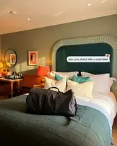 A high-quality, inviting photograph of a hotel room interior, featuring a neatly made bed with a distinctive green headboard and a travel bag placed in the foreground, creating an aspirational 'check-in' aesthetic.