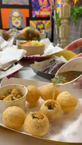 A montage of various dishes including loaded fries, golgappe, and naan, set within a vibrant, colorful restaurant space featuring a tuk-tuk.