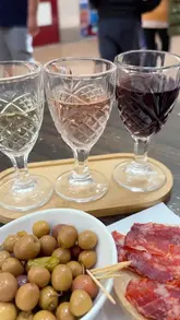A casual, eye-level shot of a wine flight and accompanying snacks, including olives and charcuterie, served on a wooden board at a market-based deli.