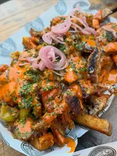 A close-up, high-angle shot of a portion of loaded fries topped with grilled chicken, melted cheese sauce, and pickled red onions, served in a branded paper-lined basket. The image highlights the textures and indulgent nature of the street food dish.