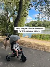 A bright, well-composed shot of a child on a tricycle positioned in the foreground of a scenic park setting by a lake. The image is used to promote the location as a destination for summer outings.