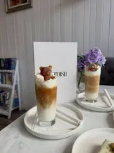 A bright, well-composed shot of two iced lattes featuring cute teddy bear-shaped ice cubes, set in a cozy cafe environment.