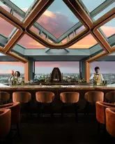 A highly polished, symmetrical shot of a bar interior featuring a glass ceiling and a sunset view over the city. The image creates an aspirational atmosphere, positioning the venue as an upscale destination for evening drinks.