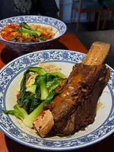 A close-up, high-angle shot of a bowl of braised short rib biang biang noodles, with a second bowl of noodles visible in the background.
