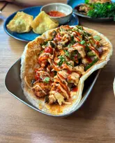 A vibrant, high-quality close-up shot of a loaded roti taco, showcasing the rich textures and sauces of the dish. The shallow depth of field keeps the focus sharp on the main subject while providing context with side dishes in the background, creating an inviting and appetising composition.