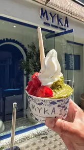 A close-up, handheld shot of a cup of Greek frozen yogurt topped with fresh strawberries and pistachio knafeh, held in front of the MYKA storefront.
