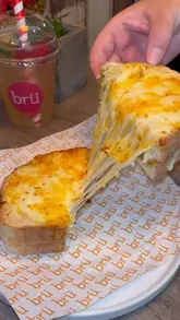 A close-up, first-person perspective shot of a grilled cheese sandwich being pulled apart to show the melted cheese, with a branded iced drink visible in the blurred background.