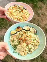A top-down, close-up shot of two bowls of prawn and zucchini pasta held by hands over a grassy background.