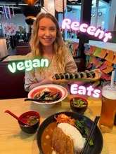 A personal, casual photo of a person holding a plate of vegan sushi, with a bowl of ramen and edamame on the table in front of them. The image is a typical 'what I ate' style post, capturing a meal out.