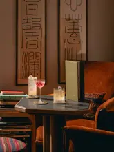 A sophisticated, atmospheric lifestyle shot showcasing two cocktails on a table within a stylish, book-filled hotel lounge setting. The image effectively captures the upscale, relaxed ambiance of the venue.