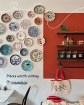 A static, well-composed shot of a cafe interior featuring a decorative plate wall and a retail shelf displaying coffee and hot chocolate products. The image serves as a visual backdrop for a listicle recommendation post about independent businesses in Edinburgh.