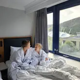 A lifestyle photograph showing two children wearing white hotel bathrobes, sitting on a bed and looking out of a large window, capturing a relaxed and comfortable hotel stay experience.