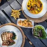 A flat-lay photograph of a pub meal served outdoors on a wooden table, featuring a chicken dish, a pork chop, fries, roasted potatoes, and a side salad.