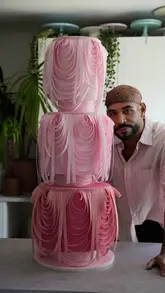 A creator demonstrates the assembly and decoration of a large, tiered pink cake using wafer paper techniques in a studio setting.