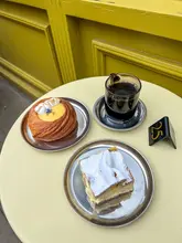 A bright, overhead flat-lay shot of a lemon curd danish, a vanilla slice, and a glass of black coffee served on metal plates at a cafe with a yellow exterior.