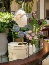 A high-quality, elegant lifestyle photograph taken inside the Corinthia London, featuring a decorative hat on a stand surrounded by lush floral arrangements in a hotel lobby setting.