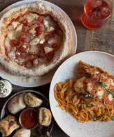 A flat-lay food photography shot featuring a spread of Italian-inspired dishes including a pizza, chicken parmigiana, and garlic dough knots, accompanied by a cocktail on a wooden table.