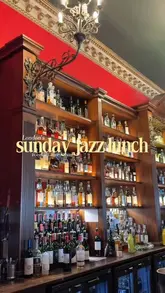 A high-quality, atmospheric shot of a well-stocked bar at Boisdale in Canary Wharf, featuring warm lighting and a classic, upscale aesthetic. The image effectively sets the scene for a Sunday jazz lunch, highlighting the venue's sophisticated interior.