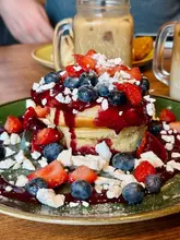 A high-quality, close-up shot of a stack of thick, fluffy pancakes topped with fresh strawberries, blueberries, crumbled meringue, and a generous drizzle of strawberry coulis, served at a brunch venue.