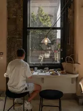 A moody, atmospheric lifestyle shot of a person sitting at a window seat in a cafe, with a view of the street outside. The scene captures a relaxed, solo moment with coffee and light bites.
