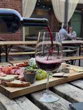 A high-quality, inviting shot capturing a wine pour over a charcuterie board on an outdoor terrace. The image effectively communicates a relaxed, social dining experience, highlighting both the drinks and the variety of food available.