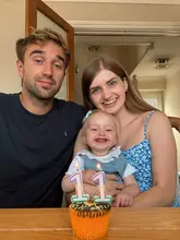 A personal, lifestyle-focused image featuring a family celebrating a monthly milestone with a cupcake. The composition is a standard portrait, with the focus placed on the subjects rather than the food item in the foreground.