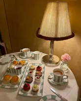 A beautifully composed, atmospheric photograph of an afternoon tea service at Claridge's. The image captures a selection of delicate pastries, scones with accompaniments, and tea, arranged elegantly on a table with warm, ambient lighting that highlights the luxury setting.