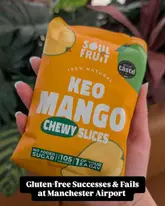 A clear, handheld product shot of a packet of Soul Fruit Keo Mango Chewy Slices, set against a soft-focus, natural background. The image effectively highlights the product packaging, serving as a simple and direct visual for a review of airport snack options.