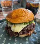 A close-up, eye-level shot of a smash burger served on branded paper, featuring a melted cheese layer and caramelized onions. The image captures the texture of the patty and the bun, though the composition is somewhat tight.